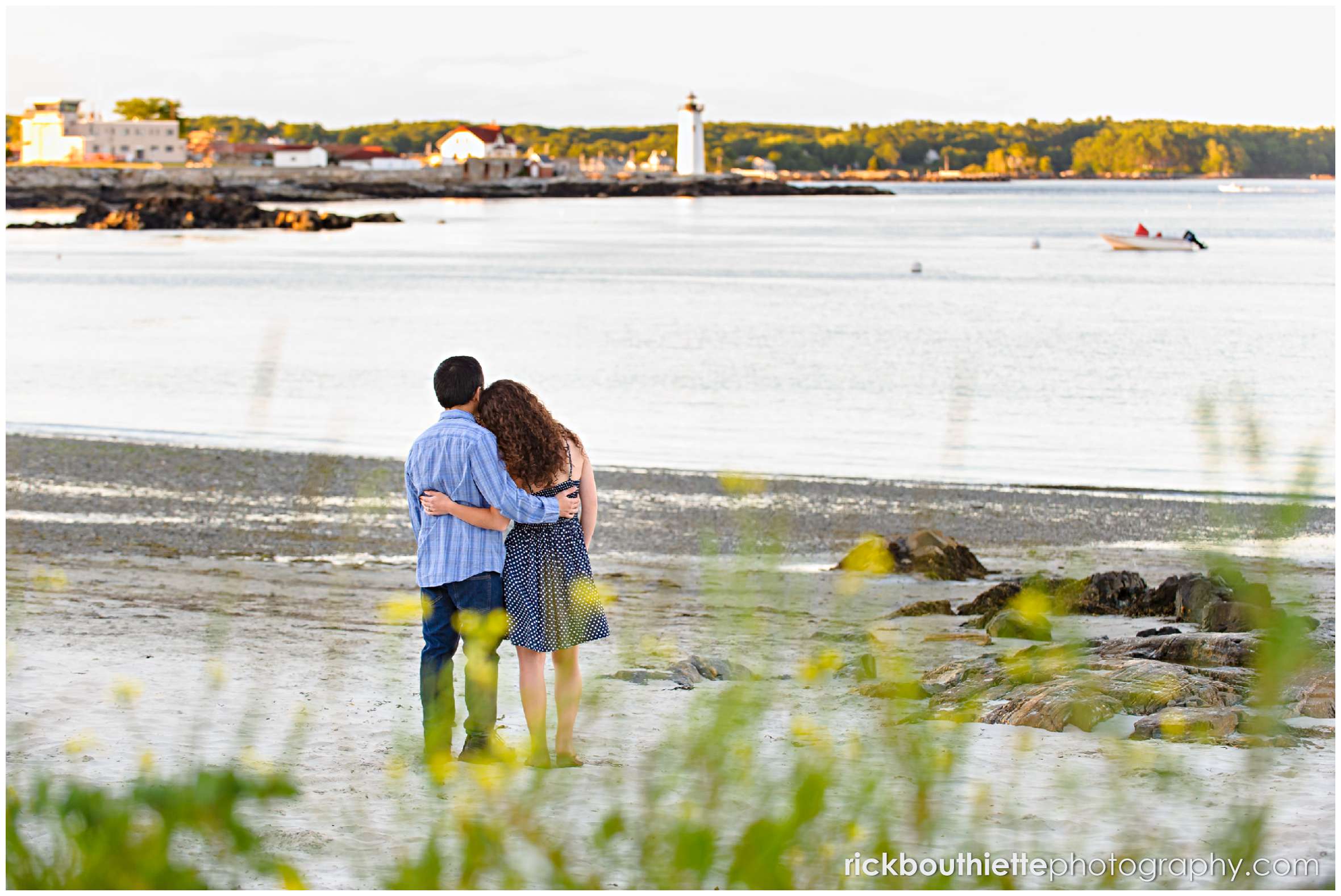 couple on the beach taking in the view of a lighthouse in the late afternoon light at their Great Island Common engagement session