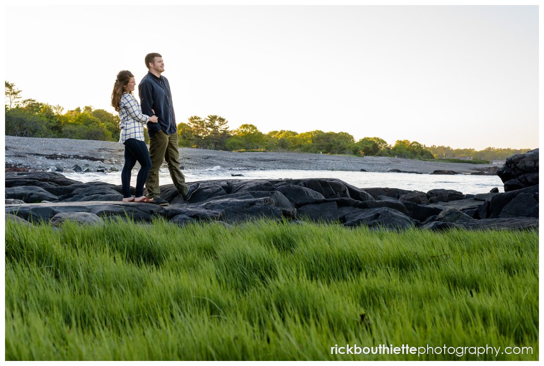 couple standing along shoreline gazing out at the ocean at seacoast engagement session